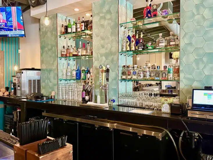 Cantina Feliz's modern, stylish bar area. The bar is framed by eye-catching mint green, hexagonal tile walls that give a sleek, contemporary vibe. Glass shelves are built into the wall and hold a wide variety of liquor bottles—mostly tequilas and other spirits, with some decorative figurines on the top shelf (like a mariachi skeleton doll and a purple skull, suggesting a playful, Day of the Dead influence). here are beer taps at the center of the bar; one tap handle is clearly marked with “Dos Equis,” indicating Mexican beer is on draft. The overall feel is clean and colorful with a nod to Mexican culture, matching what you'd expect from a stylish Mexican cantina or upscale taqueria.