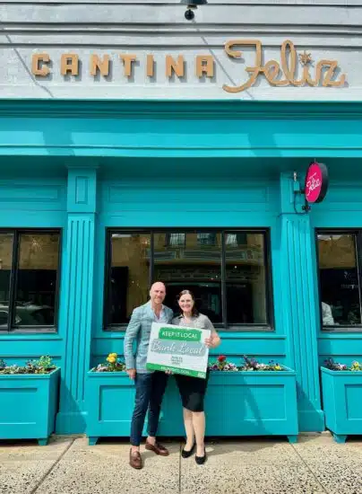 Brian Sirhal and Helen Graner standing outside of Cantina Feliz's Ambler location at 111 E. Butler Ave. Helen Graner holds a sign that says "Keep it local: bank local." The building is a striking aqua blue color and flowers in front add to the restaurant's charm.