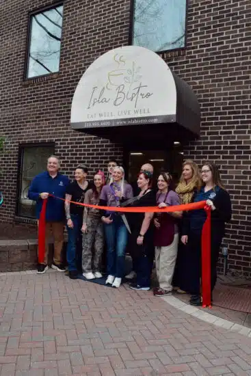 A group gathered outside of Isla Bistro with Parker Schulz for the ribbon cutting. The ribbon is red, and they are gathered in front of the brick building that is Isla Bistro.