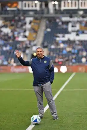 A man wearing a navy blue and gray athletic pullover stands on a soccer field, smiling and waving to the crowd. He is standing with one foot on a soccer ball, with stadium seating and fans blurred in the background.