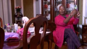 Tomika Bryant wearing all pink in living room recording herself on phone with items on table with gift basket items next to her