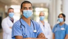 Confident multiethnic male nurse in front of his medical team looking at camera wearing face mask during covid-19 outbreak.