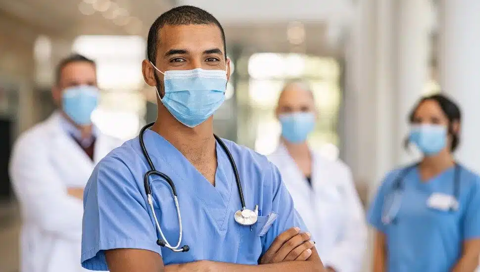Confident multiethnic male nurse in front of his medical team looking at camera wearing face mask during covid-19 outbreak.