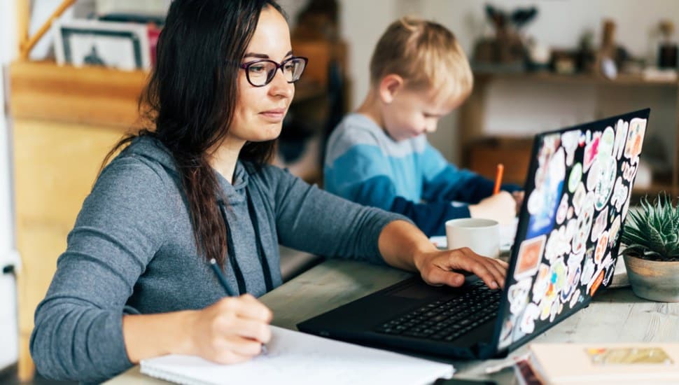 A woman looking at a laptop .computer while writing on a tablet, with her young son nearby.