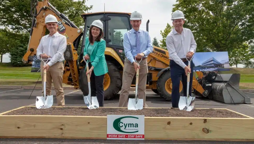 Almac Group groundbreaking, four people in construction hats breaking ground with shovel in front of excavator.