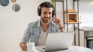 Young friendly man in good mood working as agent at call center from home while drinking coffee. Wearing headset and using laptop.