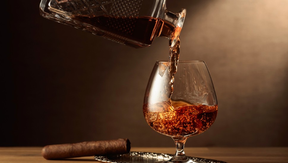 Brandy is poured from a decanter into a snifter glass. Cognac and cigar on an oak table.
