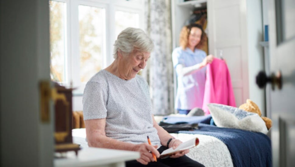 An elderly woman writing on a notepad while sitting on the bed with a nurse or aide hanging up clothes in the background.