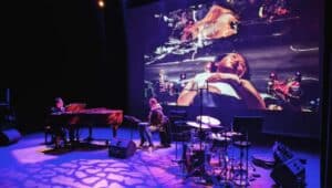 "Blind Visionaries" performance on stage man playing piano next to a set of drums near projection of visual on screen