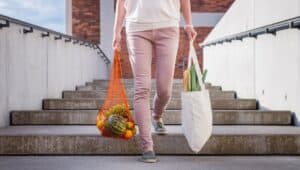 Woman walking at stairs and carrying reusable mesh bag after shopping groceries in city. Sustainable lifestyle with zero waste and plastic free