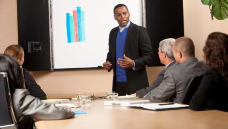 A man standing in front of a screen with a chart is guiding three men sitting at a table through a presentation.