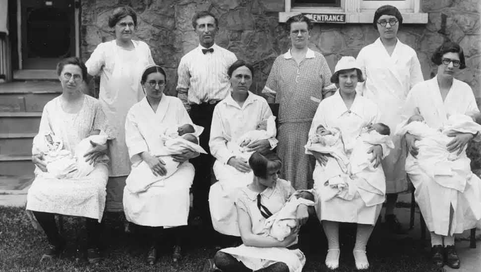 Old black-and-white photo of Mennonite women, men, and children gathered in front of a building.