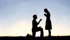A silhouette of a young man, down on one knee and holding a diamond engagement ring, proposing to his girlfriend.