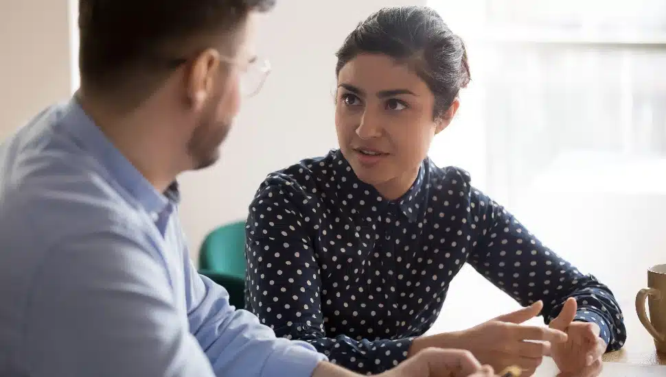 Young indian female mentor coach worker talking to male coworker teaching intern having business conversation with workmate, serious hindu manager helping colleague discussing new project in office