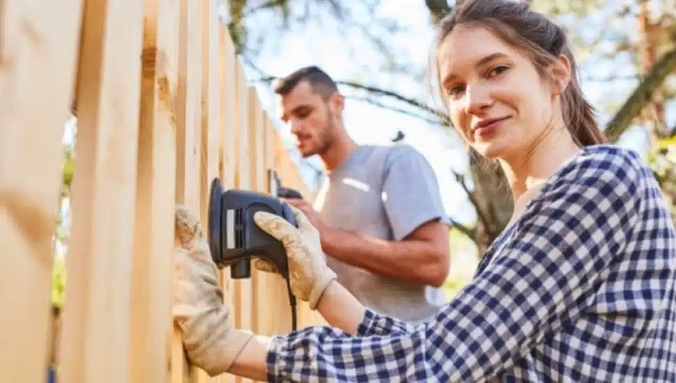 woman in gingham shirt sanding down fence wearing gloves man in gray shirt in background during day time outdoors