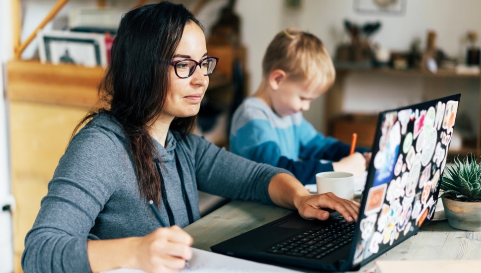 A mother doing undergraduate coursework on laptop with son in background.