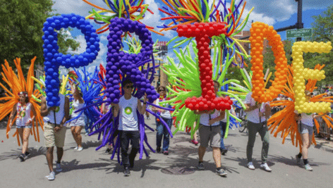 People celebrating Pride Month with a parade in the streets with balloons spelling out, "PRIDE."