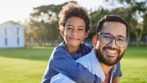 Portrait of cheerful father wearing eyeglasses giving piggyback ride to son in outdoor park at sunset. Smiling dad giving piggyback ride to his african american child at the end of school camp.