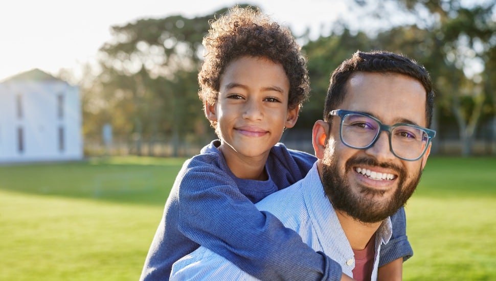 Portrait of cheerful father wearing eyeglasses giving piggyback ride to son in outdoor park at sunset. Smiling dad giving piggyback ride to his african american child at the end of school camp.