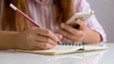 Girl With Mobile Phone Writing On Paper At Table In Classroom. Vertical view of a student texting during exam