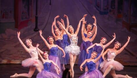 Philadelphia Dance Theatre performers in colorful costumes pose gracefully during a scene from "A Philadelphia Nutcracker," set against a festive backdrop of a snowy Victorian street.