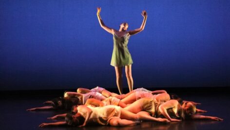 Dancers on stage at WCU. A dancer stands center stage in a powerful pose, wearing a green dress with arms extended upward, as if reaching toward the sky. She is illuminated against a deep blue background, which creates a dramatic and intense atmosphere. Surrounding her on the floor are several other dancers, lying in a circle around her, dressed in soft, neutral-colored costumes. They are positioned with their arms outstretched, creating a sense of unity and contrast with the standing figure, who appears to be the focal point of the scene. The overall effect is one of grace, strength, and harmony.