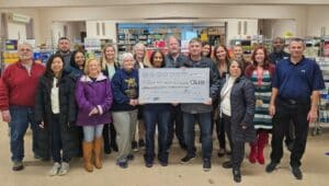 A group of individuals, including representatives from King of Prussia District, community members, and sponsors, proudly holding a large ceremonial check for $31,698 inside the Upper Merion Area Community Cupboard, surrounded by shelves stocked with food and supplies. This record-breaking donation was raised during the District's Igloo Garden on the Green event.