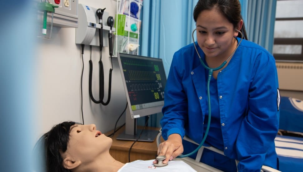 Student nurse practicing patient care using a simulation mannequin in a medical training lab, with monitoring equipment in the background.