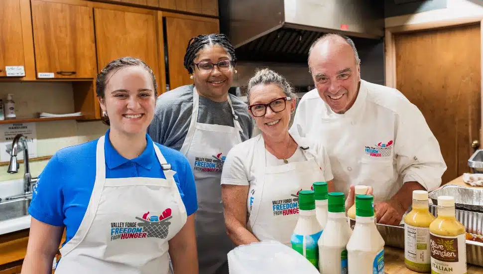 A group of four smiling volunteers wearing aprons with the logo 'Valley Forge Freedom from Hunger' stand together in a kitchen. They are surrounded by bottles of salad dressings and other food preparation items.