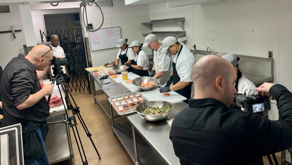 Camera crews film chefs preparing a meal.