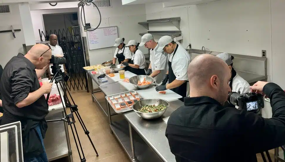 Camera crews film chefs preparing a meal.