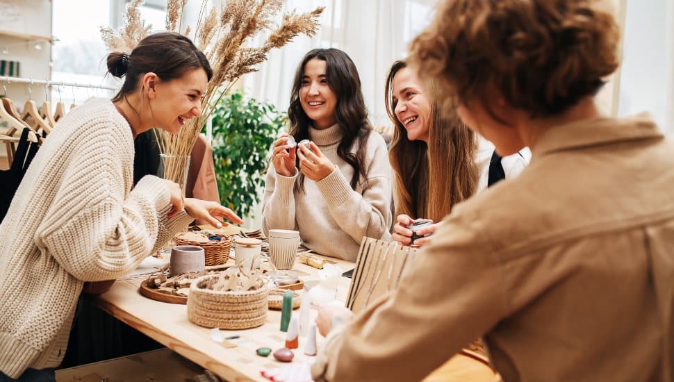Laughing friendly young women in an ecological shop picking and discussing various cosmetic. Standing in front of the table with variety of items. Talking and laughing, having a good time.