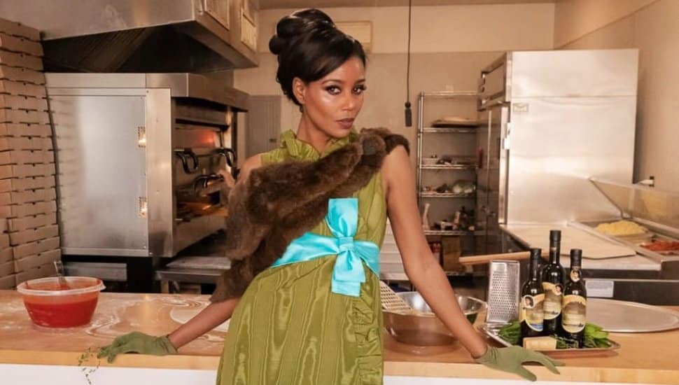 A stylish woman poses in a commercial kitchen, wearing an elegant olive-green dress with a blue ribbon, fur stole, and matching gloves. She leans against a counter with pizza-making ingredients and bottles of olive oil in the background.
