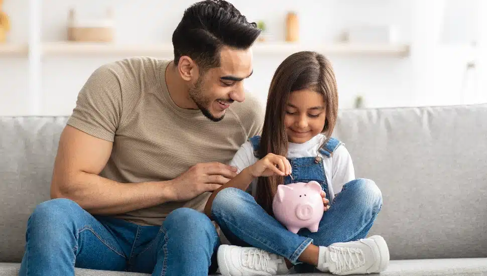 cute smiling little girl putting coin in pink piggy bank, sitting with dad on the couch at home