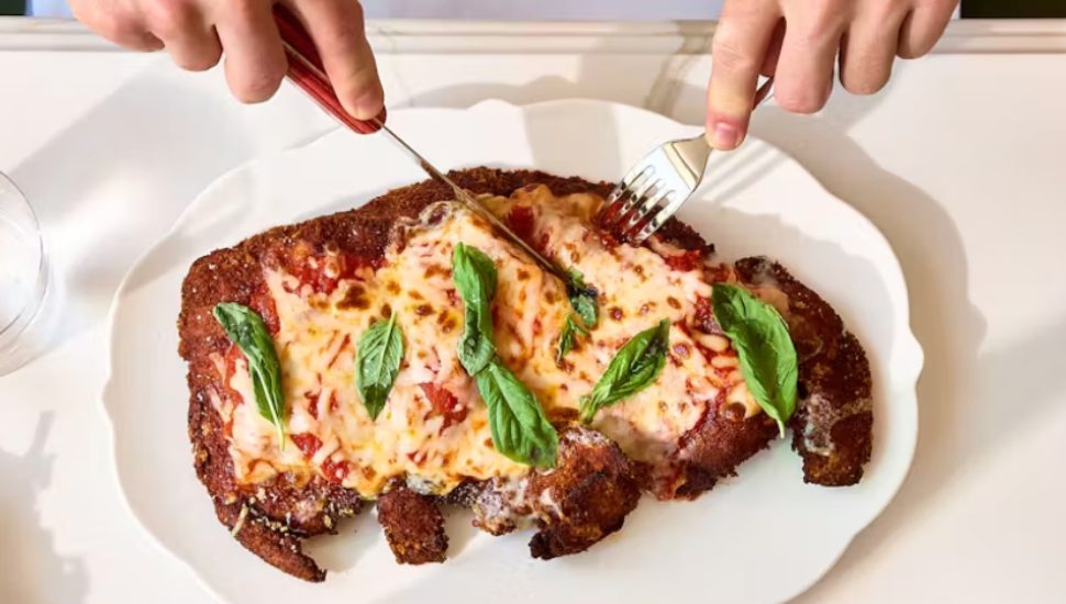 Two hands cutting into a dish of Chicken Parmesan.