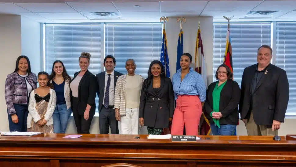 Montgomery County Commissioner Jamila Winder stands with fellow commissioners and a group of honored women entrepreneurs at a Board of Commissioners’ meeting. The group poses smiling in front of a row of flags and a large meeting table, celebrating women-owned businesses and their contributions to the local economy.