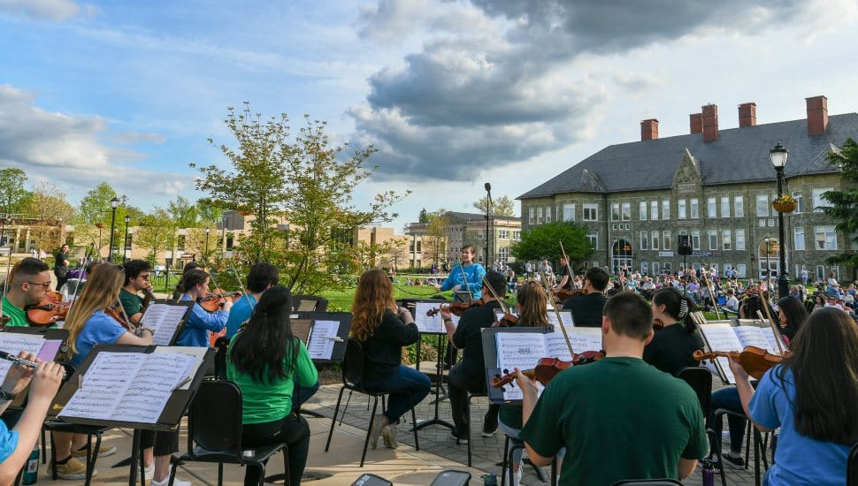 West Chester University students performing in an outdoor orchestral concert during the annual Concert on the Quad, with audience members seated on the lawn and historic campus buildings in the background under a partly cloudy sky.