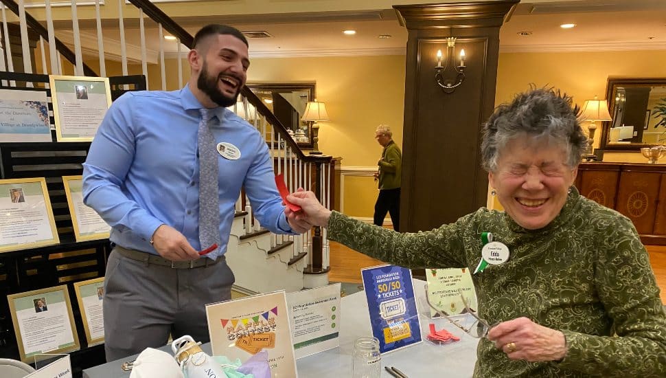 An older woman closes her eyes as she takes a chance on a 50/50 raffle ticket sold by a man standing behind a table.