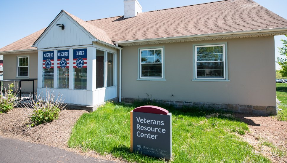 The Veterans Resources building on the MCCC campus. It's a building with American flags in the window, and a small sign is out front indicating what the building is. It's sunny and the grass is green.