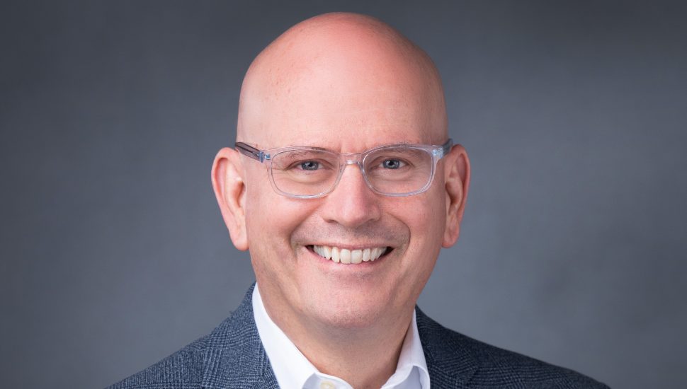 Headshot of Stephen McWilliams. He's against a gray backdrop, is bald, wears glasses, and is smiling.