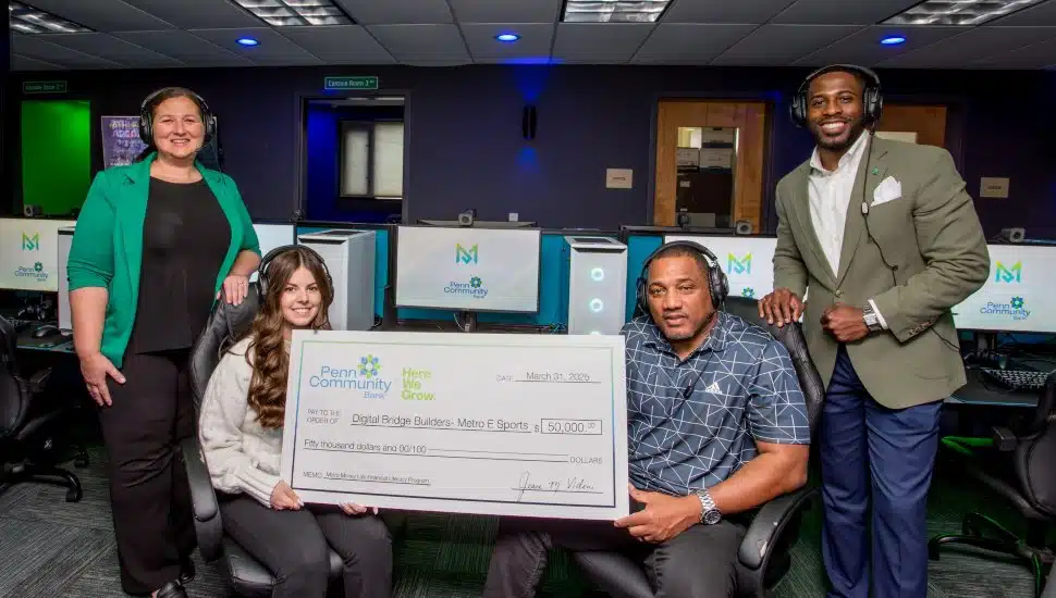 Two gamers with earphones holding a check made out to Metro Esports as part of Penn Community Bank's efforts to bring modern financial education to youth and young adults through gaming. There are also two PCB team members in the photo, for a total of 4 individuals in the photo, smiling.