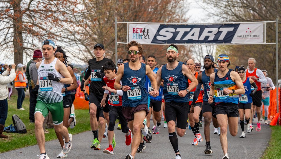 Runners take off at the starting line of the 19th annual Valley Forge Revolutionary 5-Mile Run, beneath a large banner reading "REV RUN START" with the Valley Forge logo. Participants wear race bibs and athletic gear as they begin the race on a tree-lined path, with spectators and volunteers visible in the background. The event supports Valley Forge National Historical Park.
