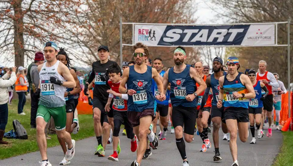 Runners take off at the starting line of the 19th annual Valley Forge Revolutionary 5-Mile Run, beneath a large banner reading "REV RUN START" with the Valley Forge logo. Participants wear race bibs and athletic gear as they begin the race on a tree-lined path, with spectators and volunteers visible in the background. The event supports Valley Forge National Historical Park.