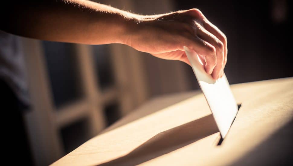 A hand dops a mail-in ballot into a drop box.