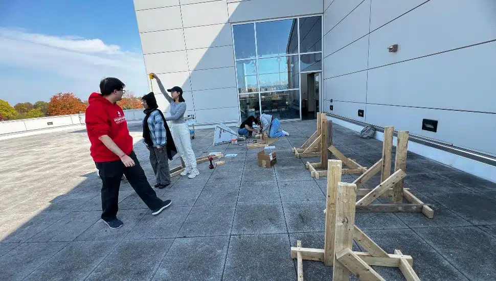 Three college students— Loujain Ahmed, Ruby Armor, and Thomas Quinn—are gathered around a partially assembled horn radio antenna on a grassy patch outside a campus science building. The antenna, mounted on a wooden base, has sleek metallic fins pointed toward the sky.