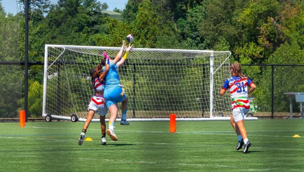 An action shot of a women's flag football game. Three women are in the photo and one is leaping to catch the ball.
