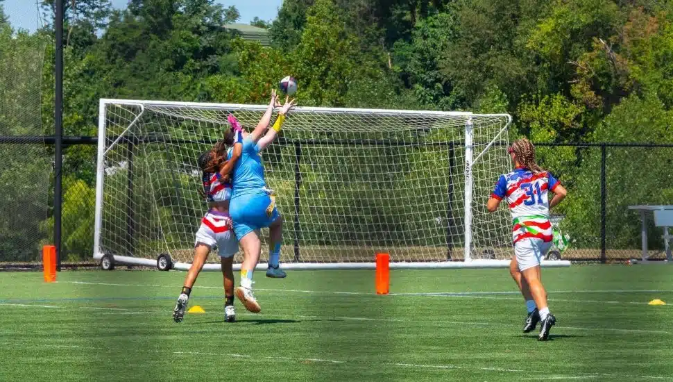 An action shot of a women's flag football game. Three women are in the photo and one is leaping to catch the ball.