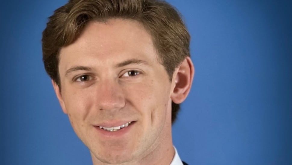 A headshot of Alex Norcross on a blue background. He has wavy brown hair and is smiling.