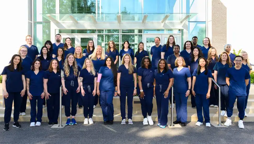 A group photo of the 38 MCCC Nursing graduates. They are wearing blue scrubs and standing outside of a campus building.