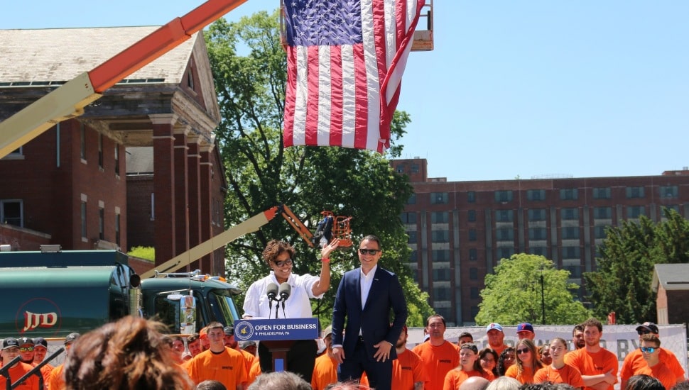 Gov. Shapiro and another official speaking at a podium in front of workers, with a crane and American flag in the background.
